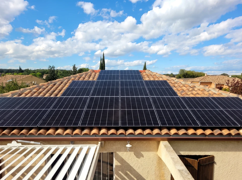 Technicien en combinaison de travail grise et jaune effectuant la maintenance d'une installation solaire photovoltaïque sur toiture, sous ciel bleu nuageux